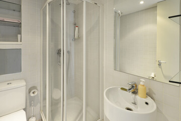 Bathroom with shower cabin with transparent screens, light marble tiling, frameless mirror and small designer sink in white porcelain, liquid soap, glass shelves and stainless steel faucets