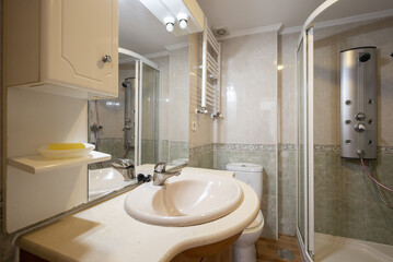 Bathroom with porcelain sink on marble countertop and wooden furniture with integrated mirror and shower cabin with water jets