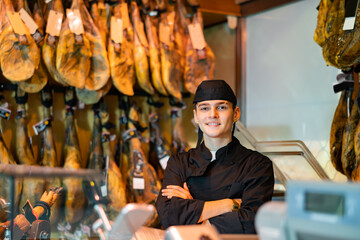 Confident smiling young butcher shop salesman in black uniform standing with crossed arms near rack with hanging jerky jamon legs behind glass counter