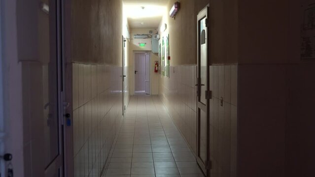 4k video. An empty corridor with no windows. There are white plastic doors in the corridor, one duty lamp is shining at the end of the emergency exit. The EXIT sign is shining green.
