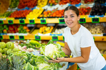 Cheerful young female in white T-shirt buying whole cauliflower during shopping in hypermarket