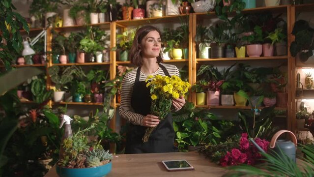 Young beautiful inspired happy woman working as a florist smelling beautiful yellow flowers wearing apron smiling. Attractive cute thoughtful female worker at florist shop enjoying spending time.