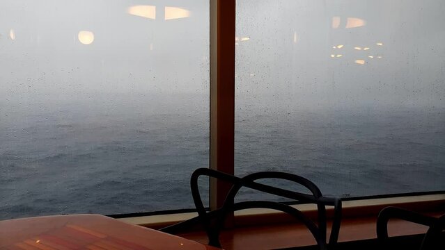 Chair and table, nobody, in front of the window of a cruise ship with the rough and rainy sea.