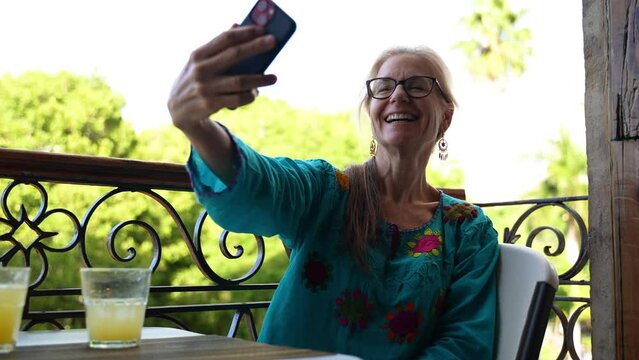 Pretty mature elderly woman with glasses taking selfies on a phone while sitting at a table on a balcony with palm trees and church in the background.