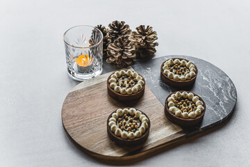 Obraz na płótnie Canvas Four tarts decorated with white cream and golden pearls placed in two lines next to each other on a board. Four cones and a candle holder with a lighten candle inside behind the board. High quality