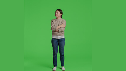 Front view of woman sitting curious and impatient in studio, posing on greenscreen backdrop. Young adult standing over isolated green screen background, being thoughtful and waiting, full body.