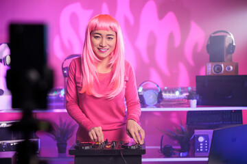 Asian dj performing techno song using mixer console while recording process with smartphone camera, putting music video on her channel for subscribers. Artist mixing at night in club