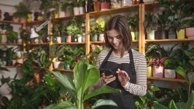 Attractive cheerful joyful young caucasian woman working as female florist using smartphone to take photo of plants at flower shop. Beautiful adult female smiling having fun working.