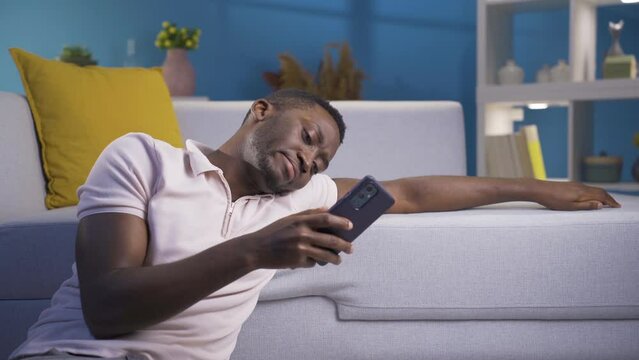 Depressed and unhappy African young man using his smartphone.
Bored lonely african young man looking at phone alone at night at home.

