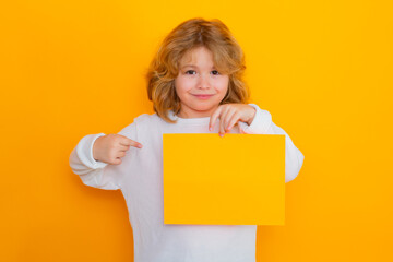 Fototapeta na wymiar Cute child showing index finger on sheet of paper, isolated on yellow background. Portrait of a kid holding a blank placard, poster.
