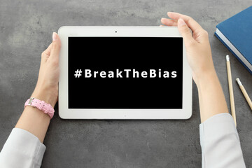 Woman holding tablet with hashtag BreakTheBias on screen at grey table, top view. Campaign theme...