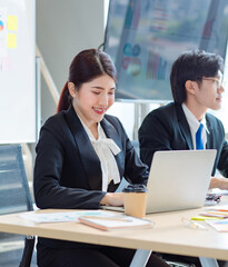 Portrait shot of Millennial Asian young professional successful female businesswoman in formal suit sitting smiling posing look at camera while male colleague working in workstation.