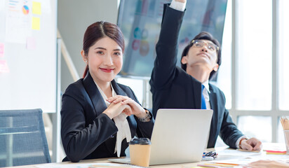 Portrait shot of Millennial Asian young professional successful female businesswoman in formal suit sitting smiling posing look at camera while male colleague working in workstation.