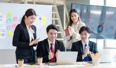 Millennial Asian young professional successful  businessman in formal suit laughing holding fists up celebrating with female and male businessman colleagues after winning job acheivement deal done