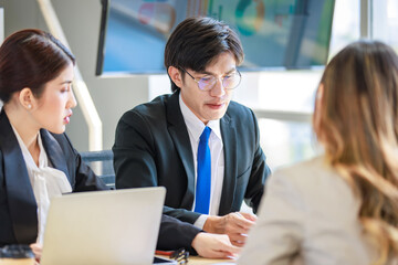 Closeup shot of Millennial Asian young professional successful male businessman employee in formal suit with eyeglasses sitting discussing brainstorming talking with female colleagues in meeting room