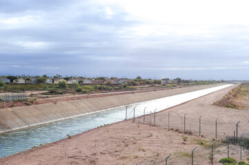 A concrete-lined water canal stretching into the distance, bordered by desert terrain, fencing, and...