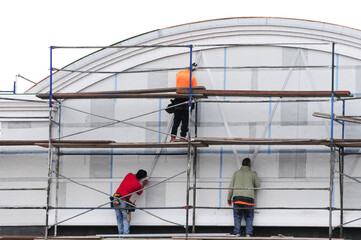 Fototapeta premium Three construction workers on scaffolding work on the curved exterior of a commercial building, performing façade installation and structural alignment tasks
