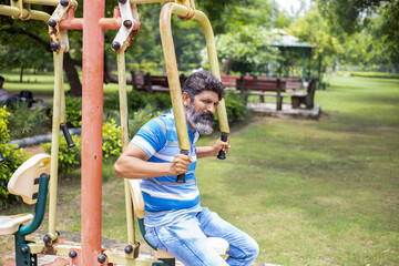 Indian senior grey bearded man exercises on the public park gym equipment outdoor.