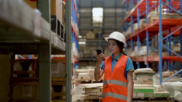 4K, Asian female clerk at large warehouse checking the number of products on shelf using a barcode scanner to scan goods. Women wear safety vests and helmets. Count the number of products in  shelves