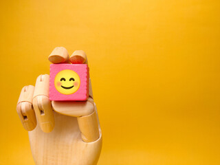 Wooden hand holding pink cube with smile icon on yellow background. Business concept.