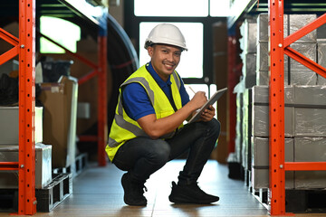 Smiling warehouse worker sitting in aisle between rows of tall shelves full of packed boxes and...