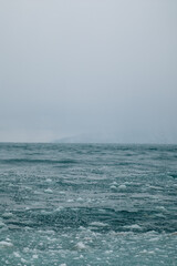 Fototapeta premium Lake shore with partially frozen banks in winter. Ice and icicles form along the edges, and the overcast sky emphasizes the cold, moody atmosphere. The scene feels serene, stark, and naturally beautif