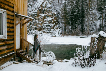 Young woman standing outdoors in winter by a mountain stream. She is dressed warmly and looks serene and contemplative. Snow-covered surroundings and flowing water create a calm, natural, and pictures