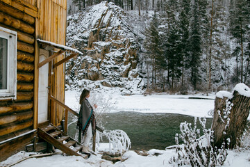 Young woman standing outdoors in winter by a mountain stream. She is dressed warmly and looks serene and contemplative. Snow-covered surroundings and flowing water create a calm, natural, and pictures