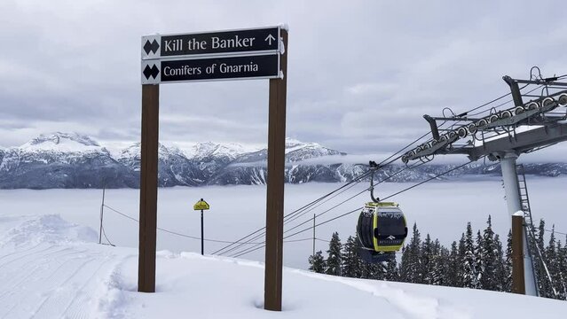 Double black diamond ski run sign on the top of Revelstoke Mt MacKenzie ski Resort with moving gondalas in the background and a view of the snowy mountains.