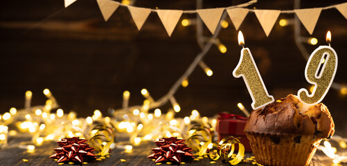 Number 19 gold burning candle in a cupcake against celebration wooden background with lights....