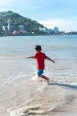 Side view happy 4 year old Asian toddler boy running playing with waves on sandy shoreline at Vung Tau beach, Vietnam