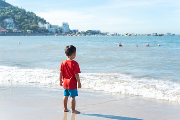 Unidentified 4 year old Asian toddler boy in red t-shirt and short standing on sandy shoreline with Vung Tau City in background