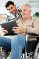man showing digital tablet to his eldery father in wheelchair - obrazy, fototapety, plakaty