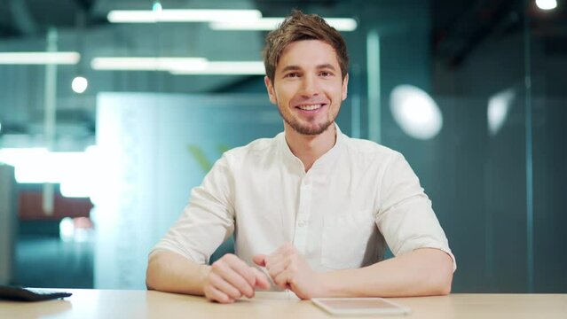 Happy smiling man sitting at office desk, looking at camera and waving hand during video call. Webcam view of handsome positive guy. He talks cheerfully with the interlocutor of the virtual meeting