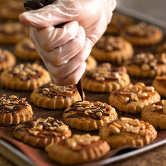 Cookies with caramel and nuts, on towel. Decorating cookies with chocolate topping. Work with pastry sack. Hand of pastry cook in glove. Serving and serve of cookies.