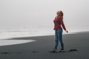 Joyful woman on empty black beach scenic photography. Picture of person with stormy sea on background. High quality wallpaper. Photo concept for ads, travel blog, magazine, article