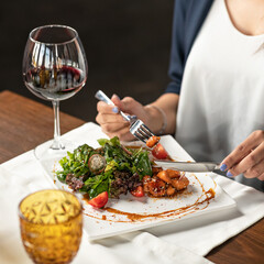 Woman is having salad and glass of red wine for lunch. Knife and fork in hands of woman. Lunch at table in restaurant. Vegetable salad on white plate onawooden table. Soft focus. 