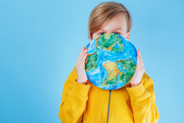 A child closing his face with a painting planet Earth, eyes looking at the camera on a blue background. Save go earth, saving environment, save clean planet, ecology concept. Sustainability project. 