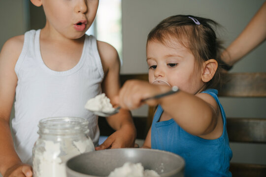 Baby Daughter Pouring A Spoonful Of Flour, Adding Ingredients While Preparing Dough For Cookies With Mother And Brother At Home. Cooking Together At Home