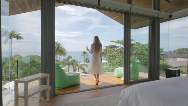 Woman Walks Through The Bedroom With Drink In The Hand To The Balcony With Sea View