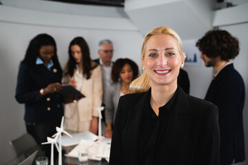 Portrait of young attractive blonde businesswoman standing and smiling. Defocused teamwork in workplace.