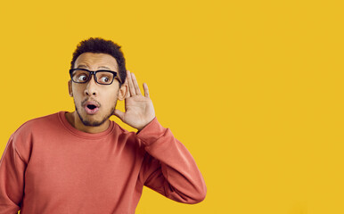 Young African American man isolated on yellow background hold hand at ear listen to news. Shocked black guy hear sale or promotion deal or offer announcement. Copy space, blank ad spot.