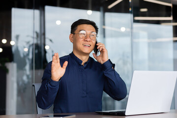 Successful and cheerful asian businessman talking on the phone with clients inside the office,...
