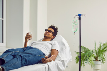 Fototapeta na wymiar African American man getting treatment at the clinic or hospital, lying on a medical bed, receiving a medication infusion through an intravenous drip, holding a glass of water and taking medicine