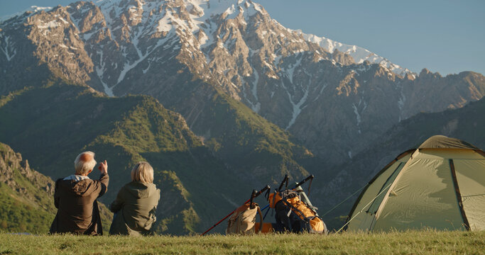 Happy senior adult mature classy couple sitting at the top of mountains, thinking of good future. Carefree mid age old husband embracing wife looking away dreaming, enjoying well being and love 