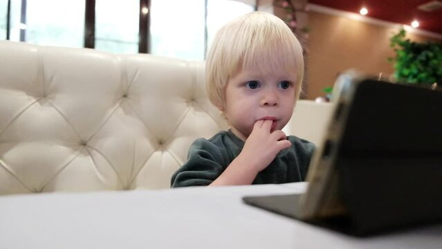 Little cute boy sits in a cafe and looks at the smartphone screen