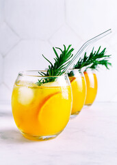 Fresh citrus juice in glasses standing in a row. Cold summer drink with citrus slices, ice and rosemary branch on white background. Close up. Selective focus. Blurred background.