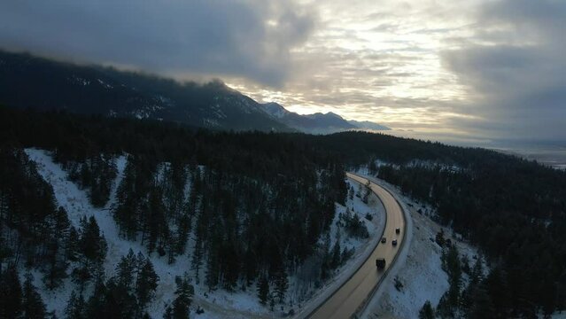 Early Morning Sunrise Aerial Reveal of Cars Travelling Along Snow-Covered Mountain Cariboo Highway 95 with Reflections on Road in Mystical, Cloudy Scene in British Columbia, Canada"