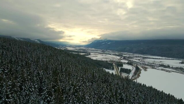 Golden Hour Aerial View of Snowy Forest and Cloud-Covered Mountain Scenery in British Columbia, Canada: Stunning Winter Landscape from Drone's Perspective