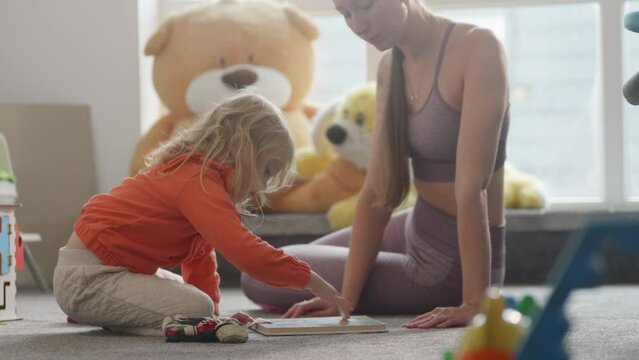 Young mother in sport outfit teaches daughter with a book. Cute blond kid, pretty women wearing bright clothes, nursury room with toys. Trying to find time for sports being a parent. High quality 4k